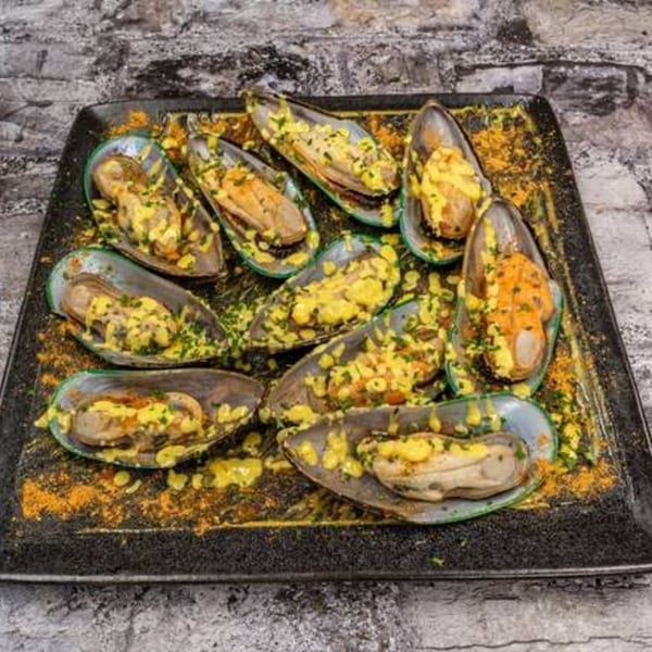 A black rectangular platter filled with green-lipped mussels topped with yellow sauce and green herbs.